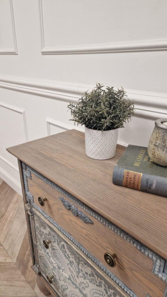 Komoda w stylu French Vintage z detalem drewnianego blatu, na którym stoją dekoracyjne książki i doniczka.