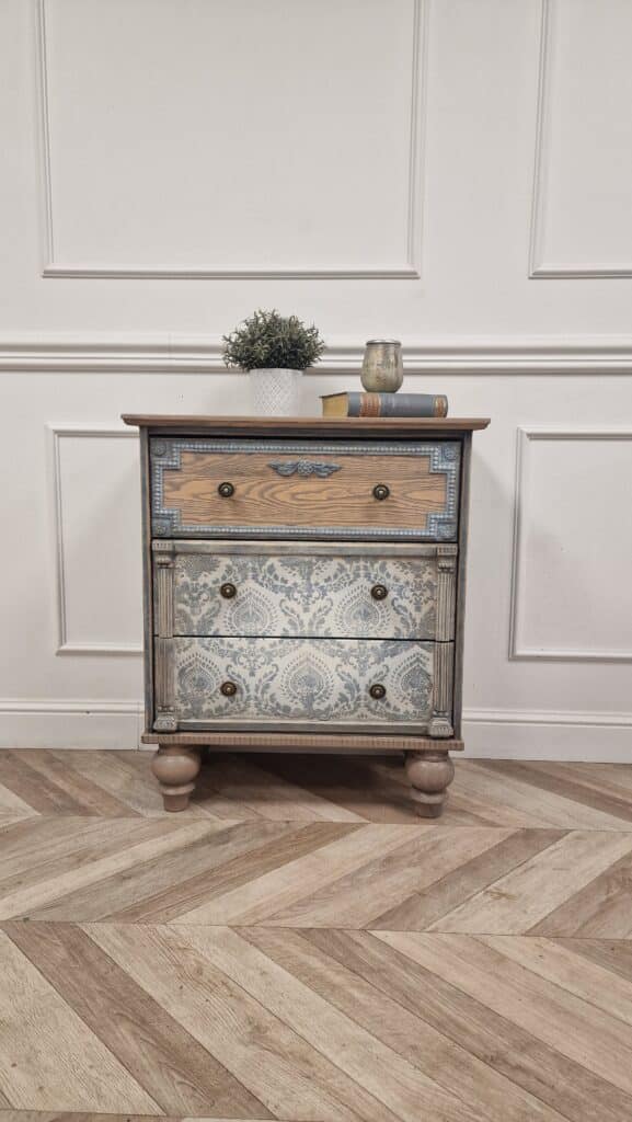 Komoda w stylu French Vintage na tle białej ściany, ozdobiona strukturalnymi wzorami i przecieranym drewnem.