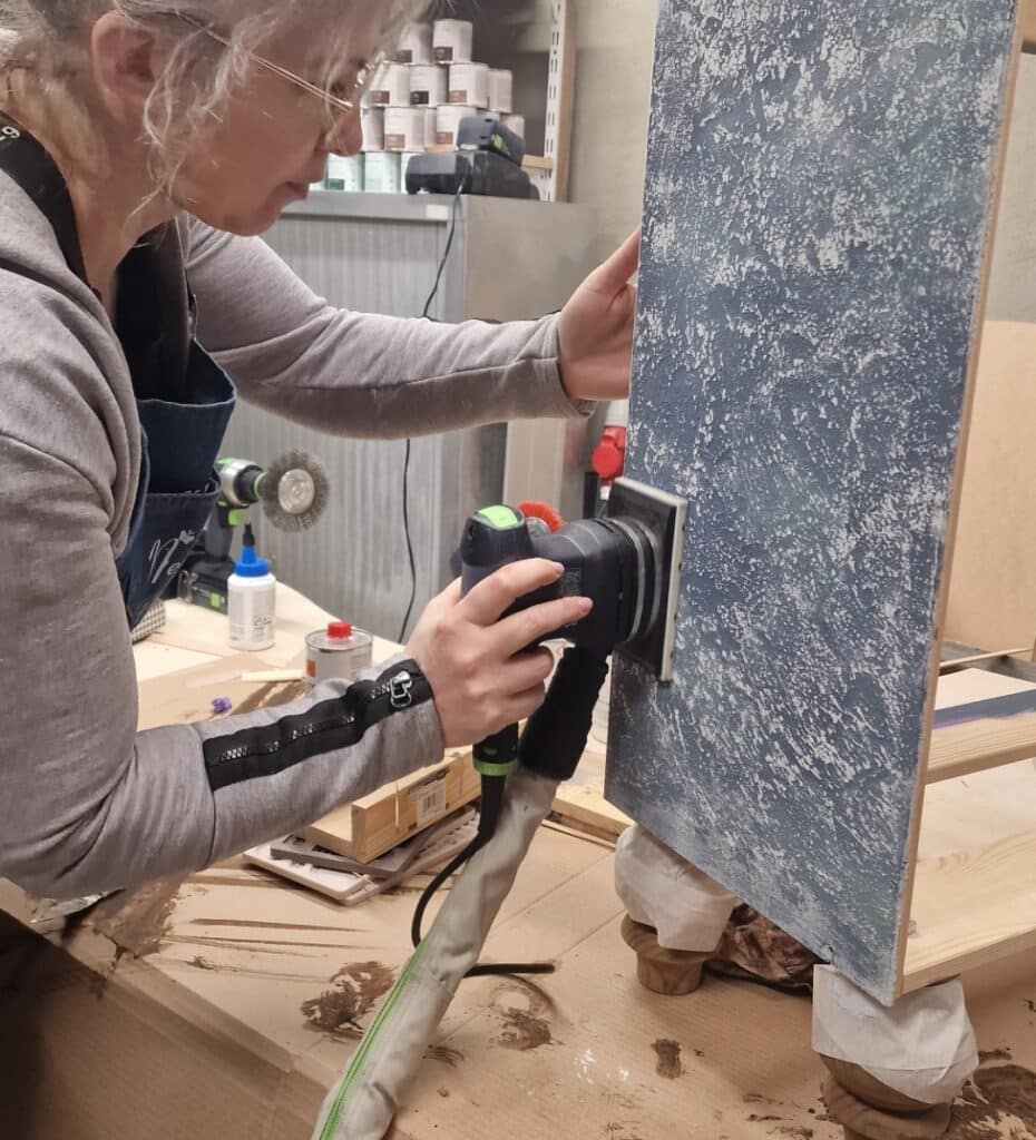 Kobieta szlifująca boczny panel komody w stylu French Vintage przy użyciu szlifierki oscylacyjnej w warsztacie stolarskim.